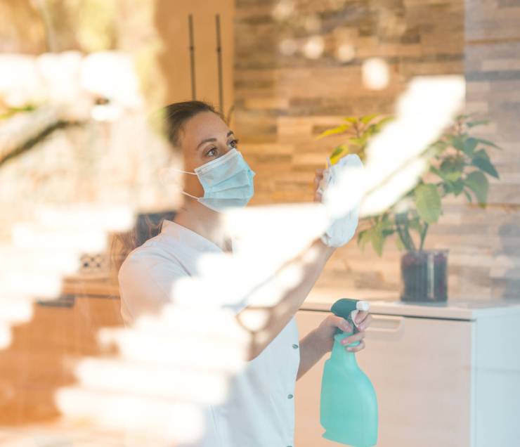 Through glass of female worker in medical mask wiping window and using spay bottle of detergent while disinfecting medical clinic during coronavirus pandemic